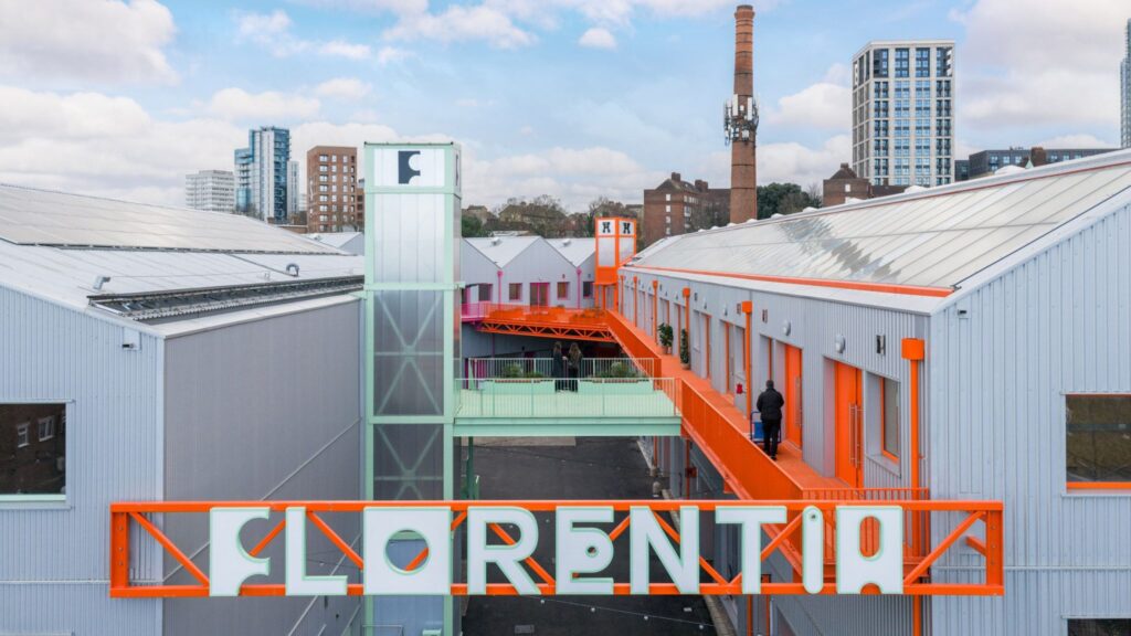 An industrial warehouse complex in London featuring light gray corrugated metal siding, vibrant orange walkways, and a large orange sign reading "FLORENTIA."