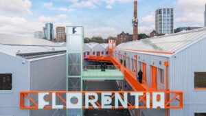 An industrial warehouse complex in London featuring light gray corrugated metal siding, vibrant orange walkways, and a large orange sign reading "FLORENTIA."
