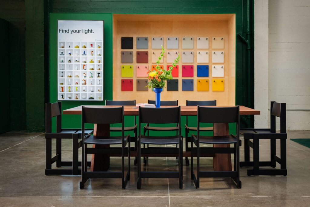 A modern showroom featuring a long wooden table with black minimalist chairs, set against a vibrant green wall with a recessed plywood display of colorful swatches and a "Find your light" product poster.