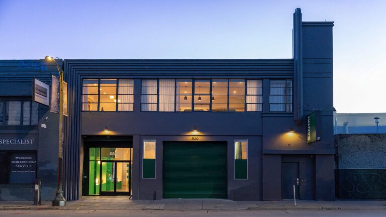 The exterior of the Gantri lighting showroom in San Francisco, featuring a dark gray industrial building with Art Deco accents, glowing windows, and a green roll-up garage door at twilight.