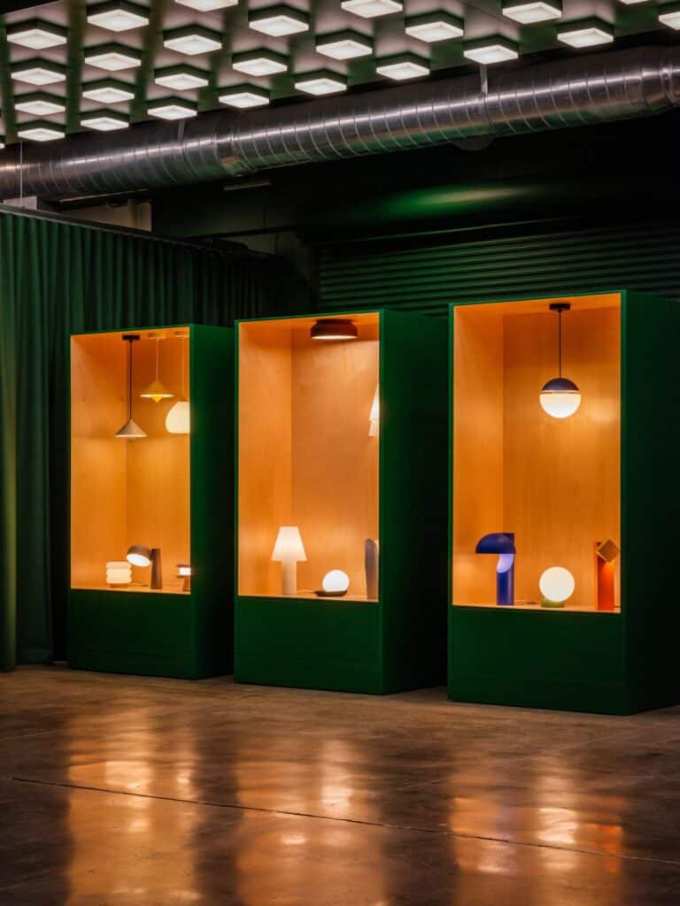 Three large forest-green rectangular display pods with warm wood interiors showcasing various modern 3D-printed lamps in a showroom.