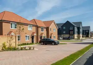 A view of a modern housing development featuring red brick semi-detached houses with orange tiled roofs and dark grey multi-story apartment buildings under a clear blue sky.