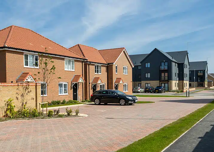 A view of a modern housing development featuring red brick semi-detached houses with orange tiled roofs and dark grey multi-story apartment buildings under a clear blue sky.