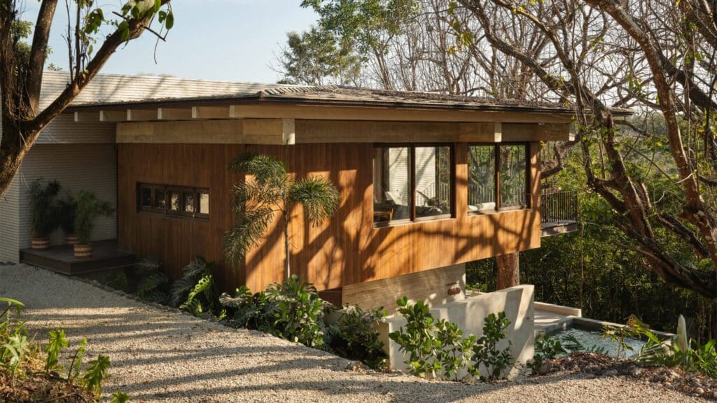 An elevated view of a modern wooden home featuring large windows and a flat roof, overlooking a private pool and dense tropical forest.