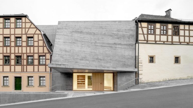 A modern gray board-marked concrete building extension nestled between two traditional German half-timbered houses on a sloping street.