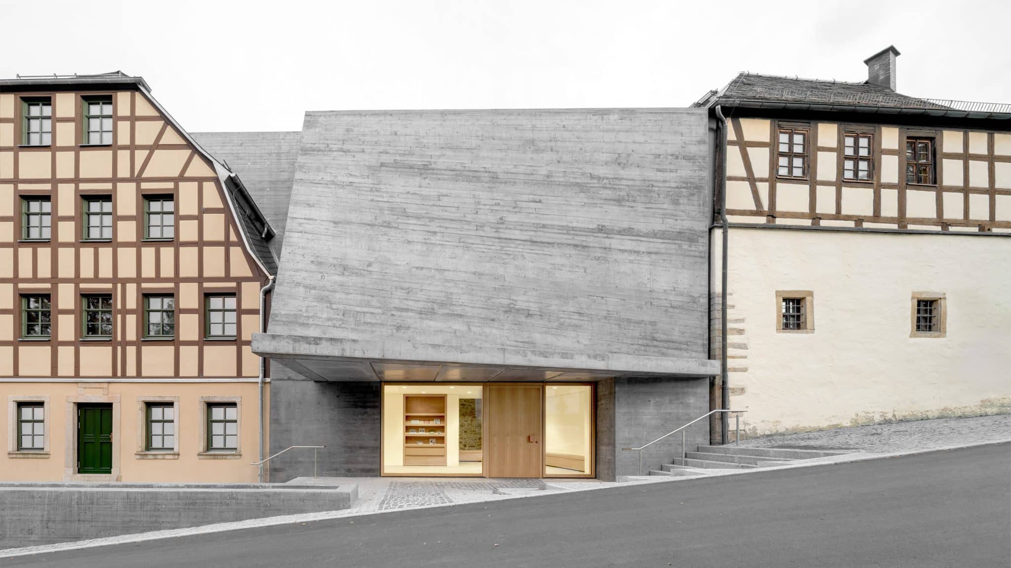 A modern gray board-marked concrete building extension nestled between two traditional German half-timbered houses on a sloping street.