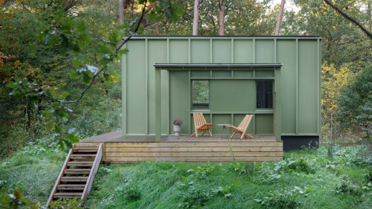 A modern, sage-green minimalist cabin with vertical wood paneling, featuring a raised wooden deck with two lounge chairs and a small table, nestled within a lush green forest in the Netherlands.