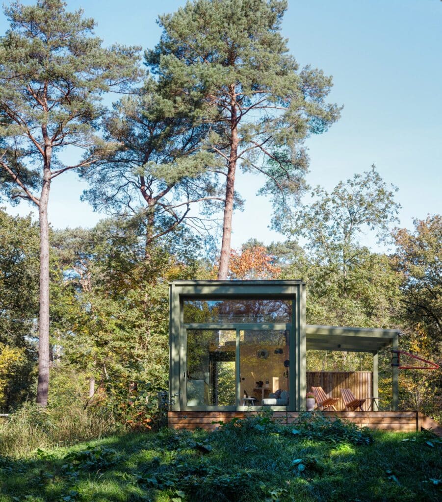 A wide exterior shot of a small, olive green modern cabin with a large glass facade and an adjacent covered wooden deck, situated in a dense forest with tall pine trees under a blue sky.