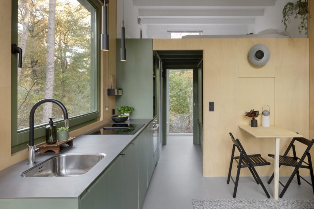 Interior of a compact wooden cabin showing a sage green kitchenette, a light plywood wall with a fold-out dining table, and a lofted sleeping area above.