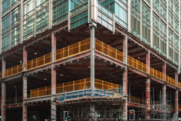 A corner view of a high-rise building under construction, showing a glass facade on the upper floors and an exposed steel structural frame with yellow safety railings on the lower levels.
