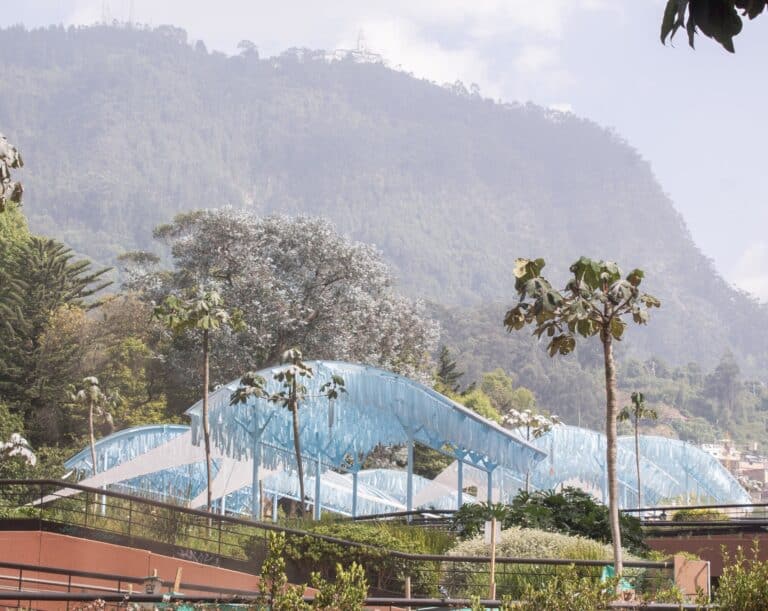 A side view of the "River Memory" ephemeral structure with blue fabric strips under a translucent roof, set against a backdrop of a lush green mountain in Bogota.