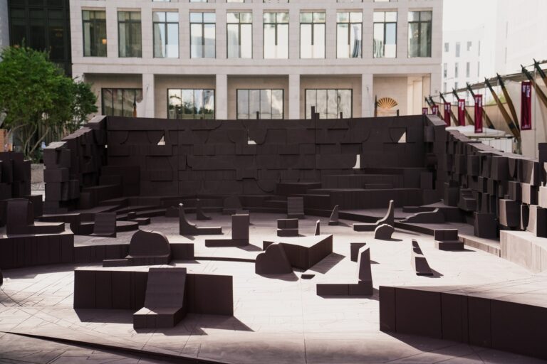 A wide interior view of the architectural installation "In the Assembly of Loved Ones" by Counterspace, featuring dark brown modular wooden elements arranged in a communal gathering space.