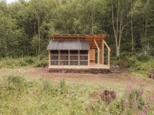 Exterior view of GLØDE shelter in Beiarn, Norway, featuring recycled wood siding and a corrugated metal roof against a lush forest background.