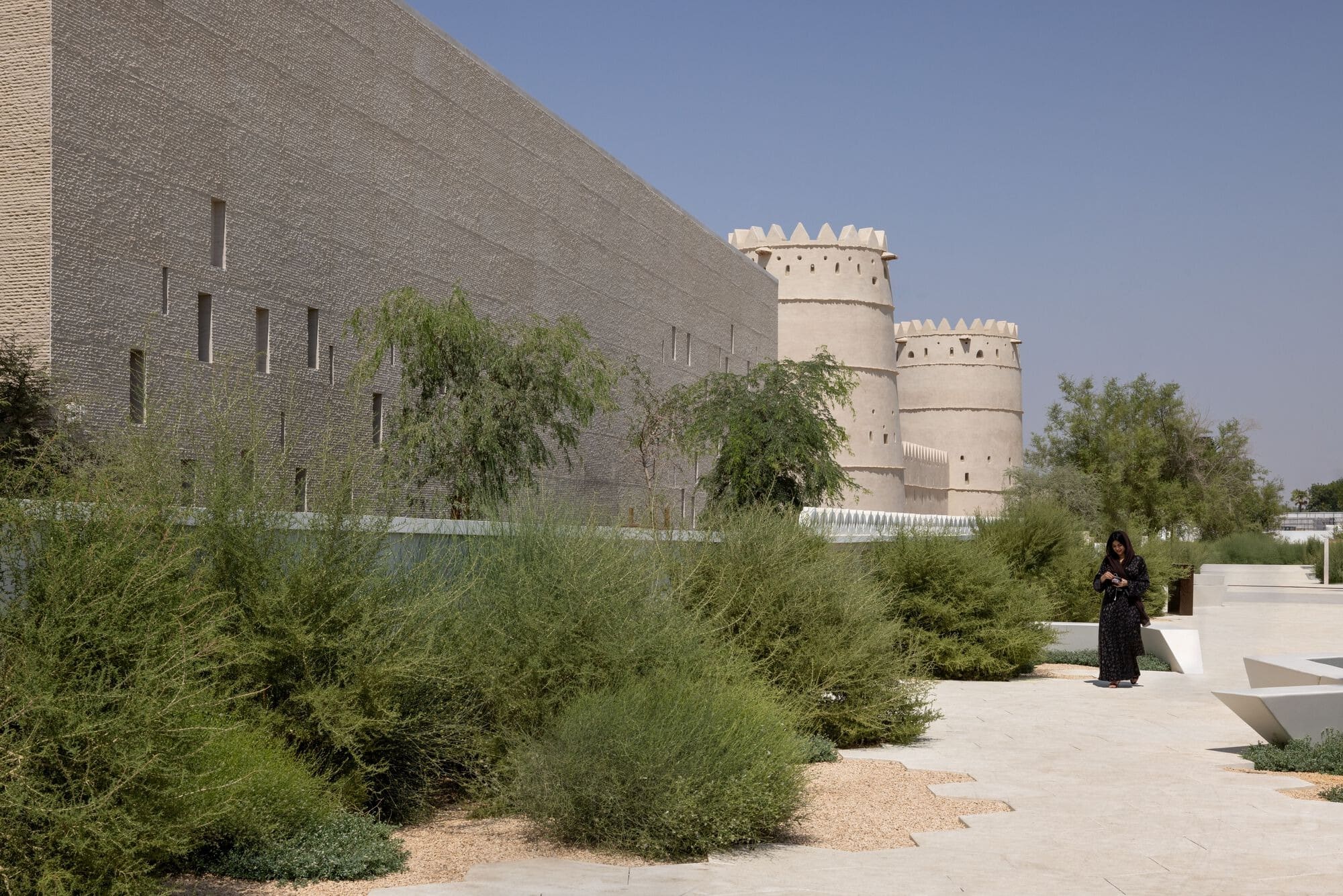 Contemporary Al Ain Museum stone facade next to the historic Sultan Bin Zayed Al Nahyan Fort towers and desert landscaping.