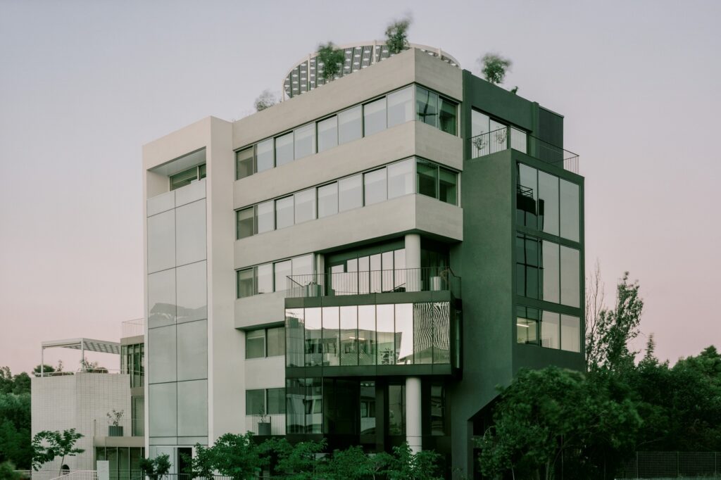 A contemporary office building rehabilitation showing a multi-textured facade with glass panels, grey concrete walls, and a rooftop garden under a clear sky.