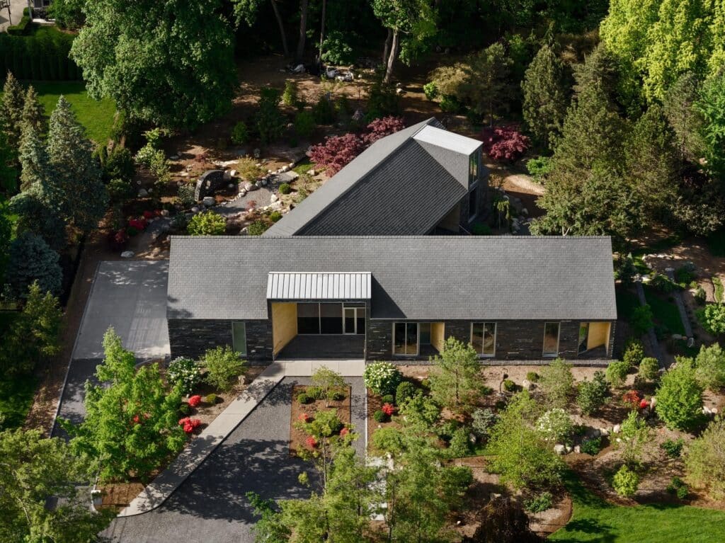 Aerial drone view of a Y-shaped modern house with dark slate roofing and stone walls nestled in a lush, park-like forest setting.