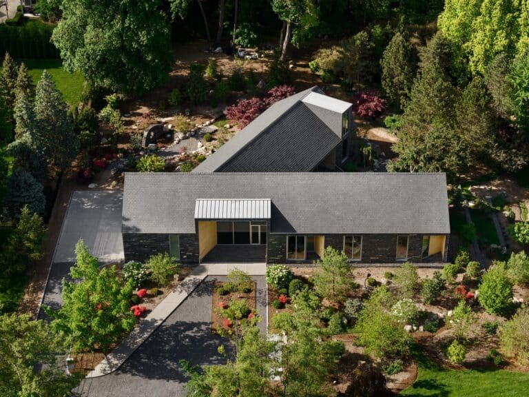 Aerial drone view of a Y-shaped modern house with dark slate roofing and stone walls nestled in a lush, park-like forest setting.
