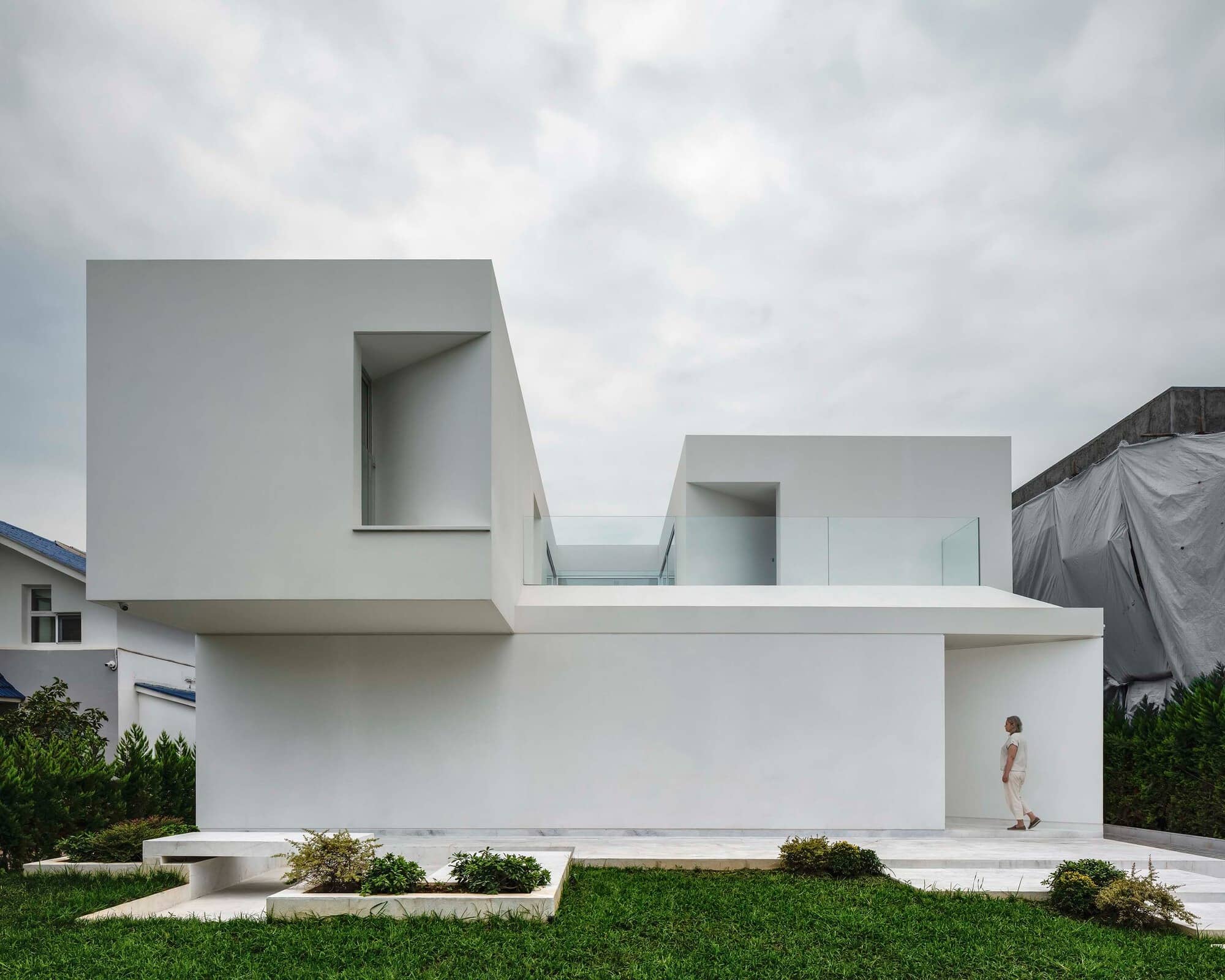 Minimalist white villa facade with geometric volumes and a central entrance, showing a woman walking on the terrace.