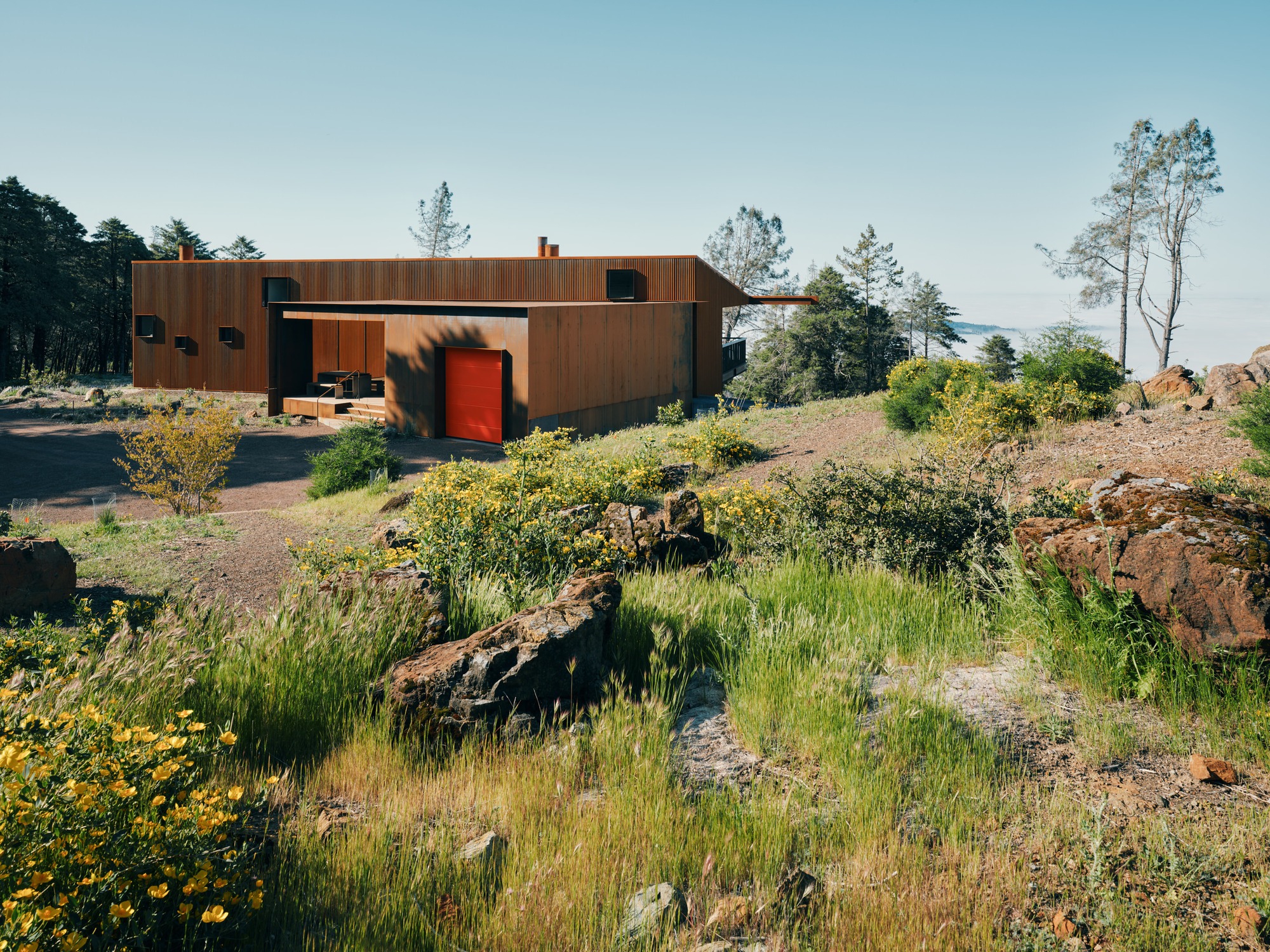Exterior view of Healdsburg house featuring fire-resistant weathered steel cladding and a bright red garage door in a mountainous landscape.