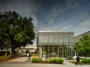 Modern glass facade of Greenhill School Valdes STEM Center with exterior sunshades and students walking in front of a large oak tree.