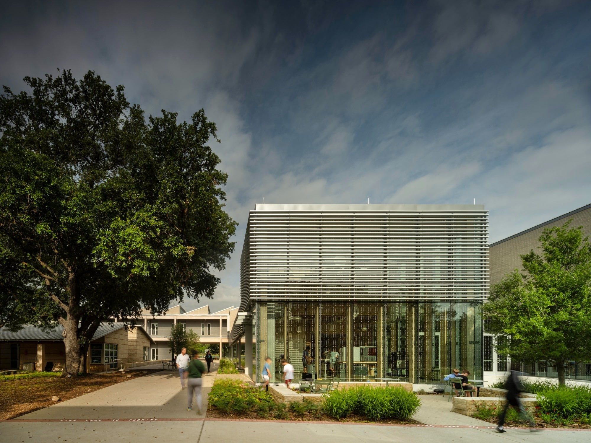 Modern glass facade of Greenhill School Valdes STEM Center with exterior sunshades and students walking in front of a large oak tree.