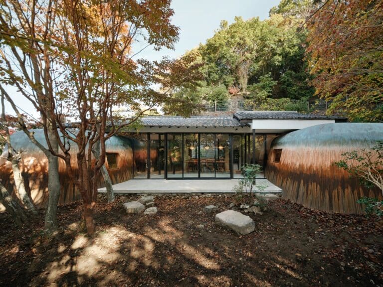 Exterior of Haniyasu House during the day showing the earthen mounds tucked under a traditional tiled roof, surrounded by autumn trees.