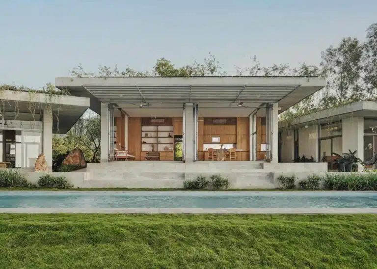 Modern hilltop house with open concrete pavilion, swimming pool, and lush green lawn under a clear sky.
