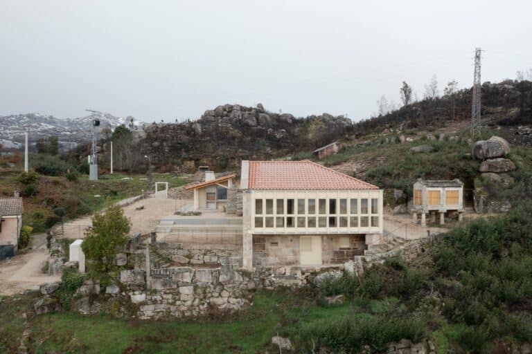 Wide aerial view of the Social Center in Moimenta showing the rehabilitated stone house, the new wooden pavilion, and a traditional Galician H&oacute;rreo on a terraced landscape.