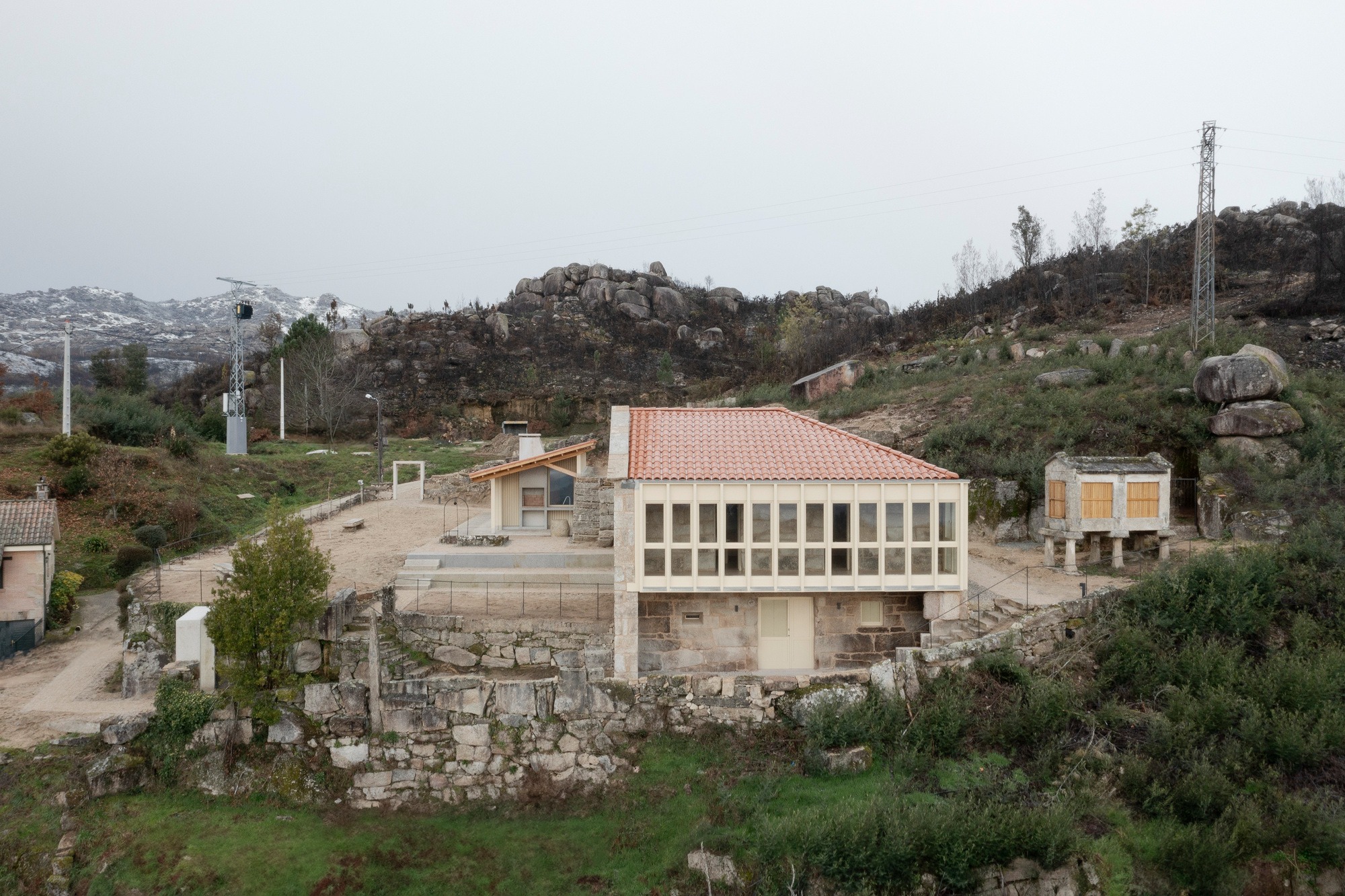 Wide aerial view of the Social Center in Moimenta showing the rehabilitated stone house, the new wooden pavilion, and a traditional Galician Hórreo on a terraced landscape.