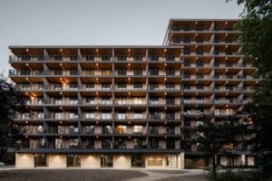 A wide dusk shot of the Valckensteyn timber residential building in Rotterdam, showing the illuminated wooden balconies and the travertine stone base.
