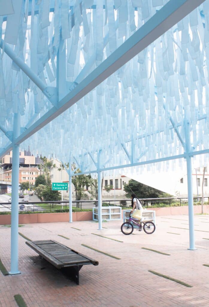Interior view of the pavilion showing thousands of blue fabric strips hanging from a light steel frame, with a person riding a bicycle in the background.