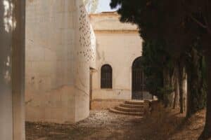 A side view of the modern concrete Requena Pantheon with small square apertures next to a traditional cemetery building with arched doors.