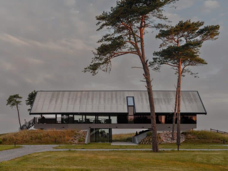 A modern coastal residence in Latvia built on elevated concrete foundations with a large gabled fiber cement roof, surrounded by tall pine trees at sunset.