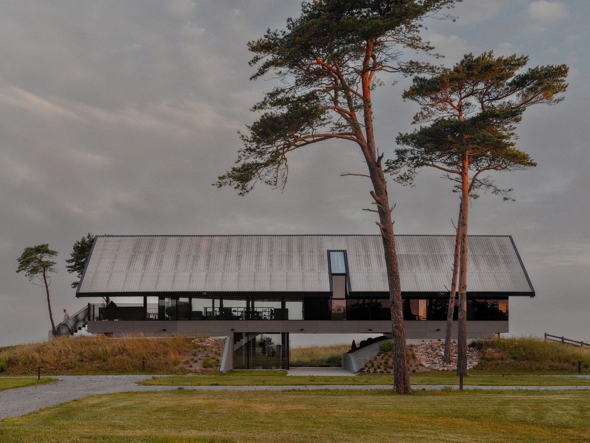 A modern coastal residence in Latvia built on elevated concrete foundations with a large gabled fiber cement roof, surrounded by tall pine trees at sunset.