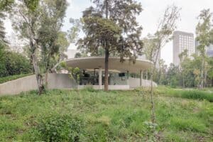 A modern circular pavilion integrated into a grassy hill at Chapultepec Park, featuring floor-to-ceiling glass walls and a thin concrete roof.