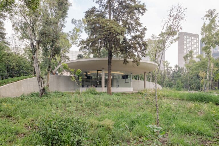 A modern circular pavilion integrated into a grassy hill at Chapultepec Park, featuring floor-to-ceiling glass walls and a thin concrete roof.