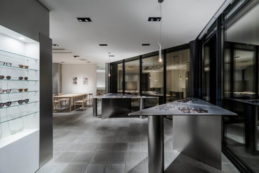 Interior view of Ashiya Optical Store showing triangular display tables and a curved glass facade overlooking the city.