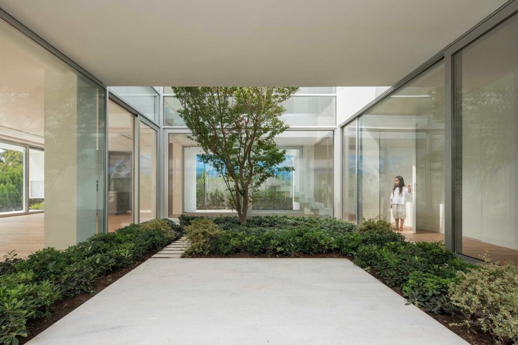 Interior courtyard of a modern villa featuring a central tree and large glass walls, with a child standing by the door.