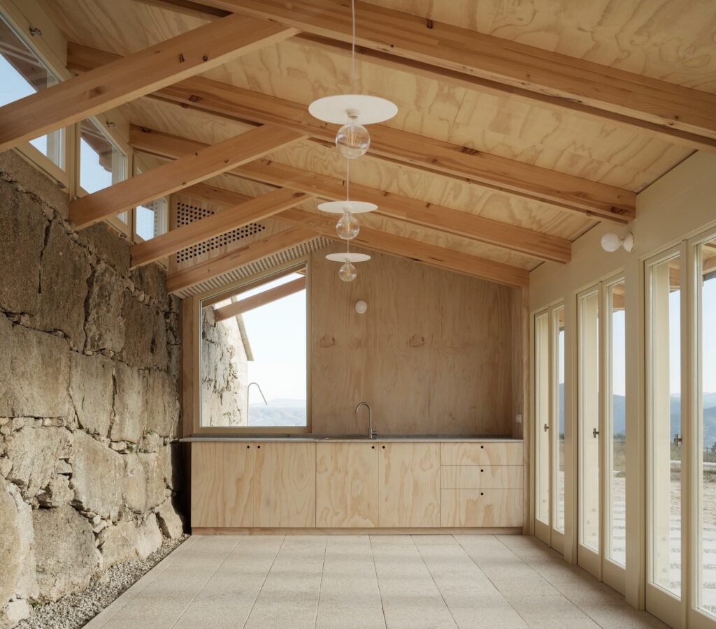 Interior view of the multi-purpose pavilion featuring a wooden gabled roof, a large stone wall, and a minimalist kitchenette with plywood finishes.