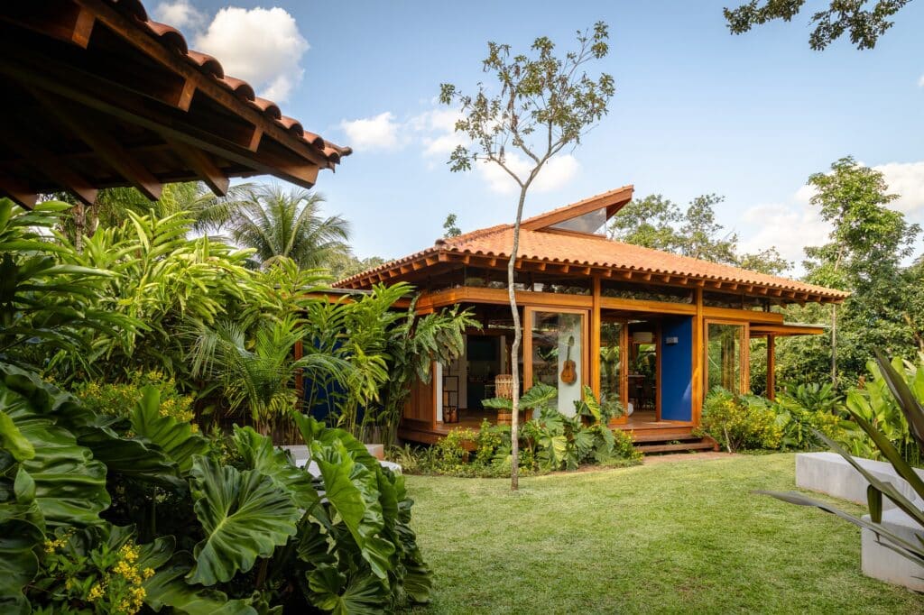 Central glass-walled pavilion with a tilted wooden roof surrounded by tropical plants and a manicured lawn.