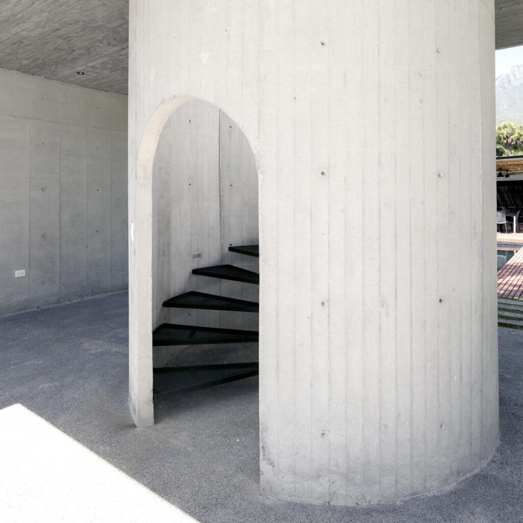 Close-up of a cylindrical concrete structure featuring a minimalist arched opening and a black metal spiral staircase.