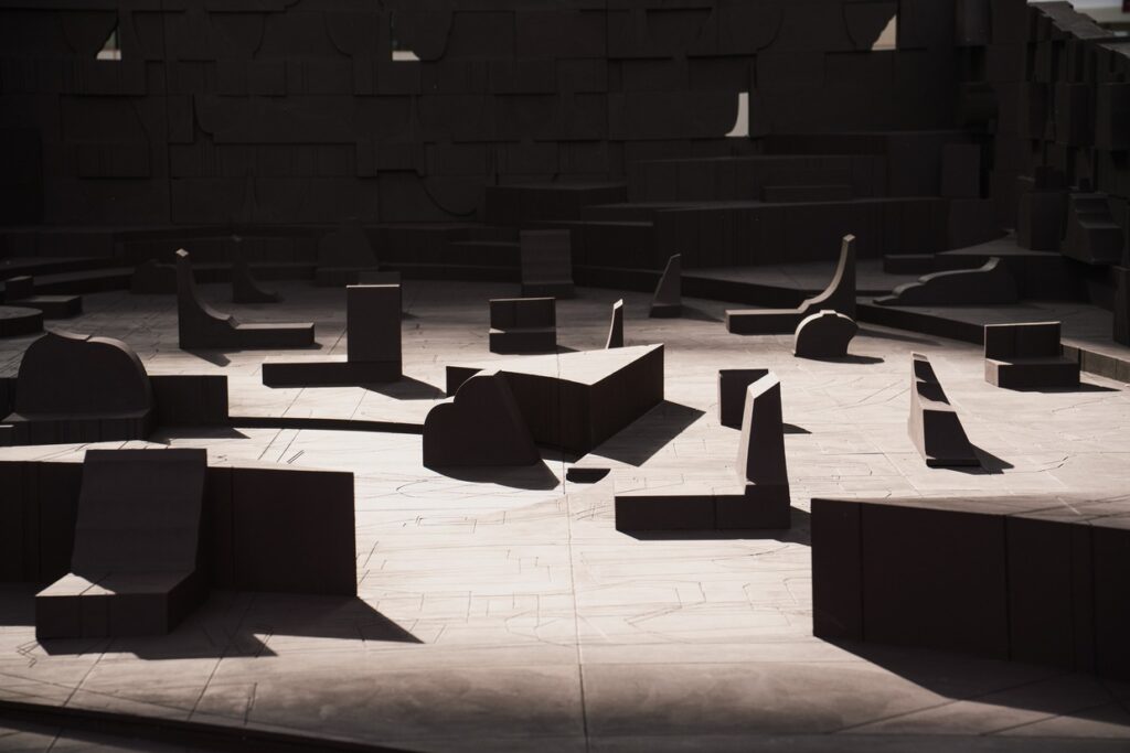 Low-angle shot of abstract wooden seating modules scattered across a floor engraved with geometric patterns, part of the "In the Assembly of Loved Ones" project.