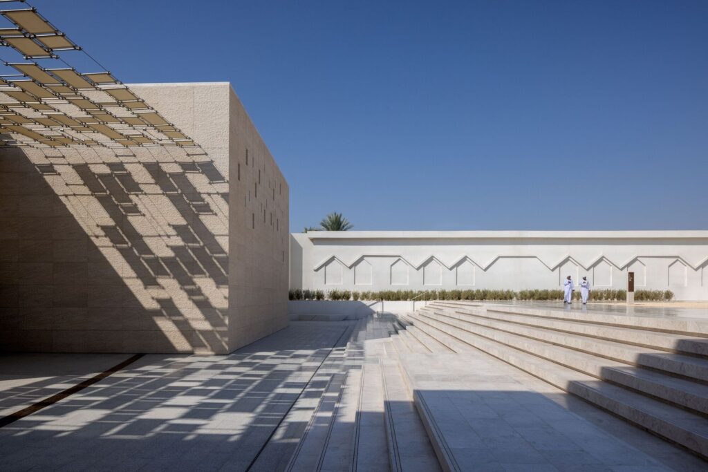 Outdoor courtyard of Al Ain Museum featuring modern geometric stone walls, grand stairs, and a decorative trellis casting shadows.