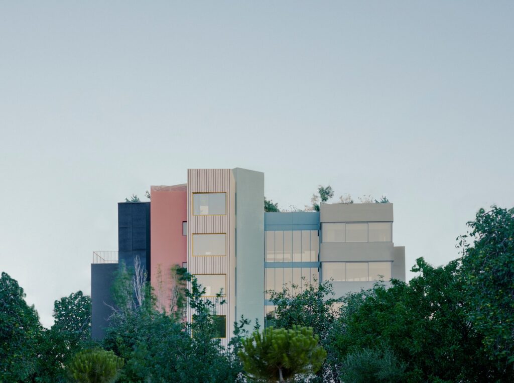 A vertical architectural shot of the building facade showing distinct color blocks in pink and pale blue contrasted against green treetops.