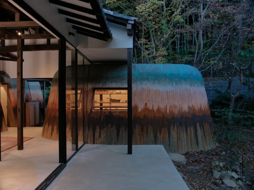 Exterior detail of an earth-textured room addition at Haniyasu House with a large glass sliding door and garden view.