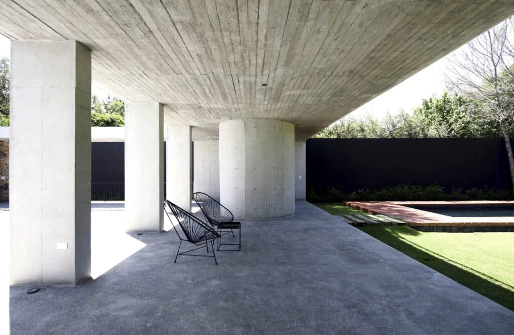 Black Acapulco-style chairs on a concrete patio under a heavy concrete roof, looking out toward a pool and dark garden wall.
