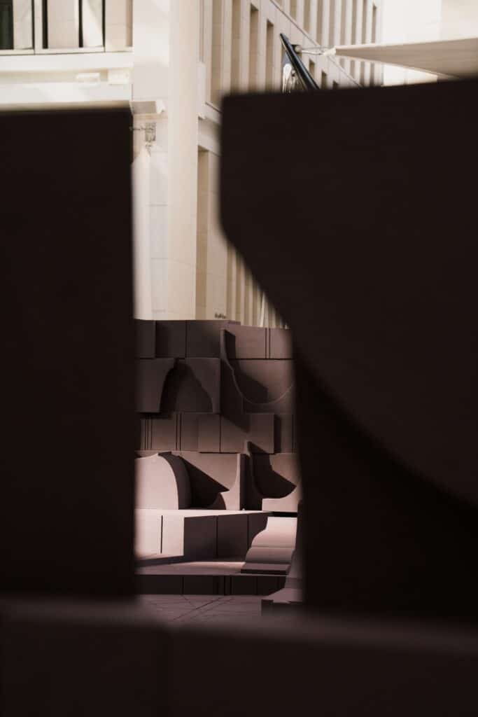 An artistic framed view through the dark wooden structures of the installation, showing light hitting the textured surfaces and background buildings.