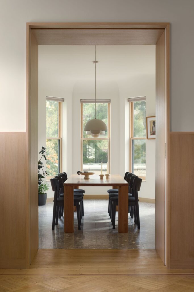 A minimalist dining room viewed through a wooden frame, featuring a long wood table and a mid-century modern pendant light.