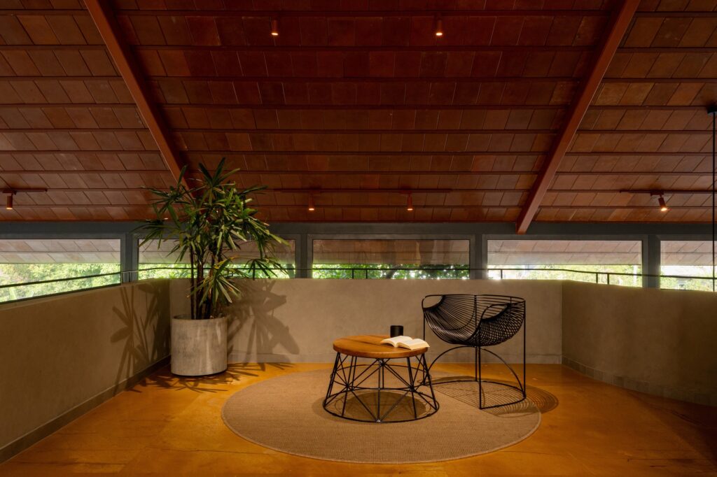First-floor mezzanine sitting area with a wooden ceiling, terracotta tiles, and a minimalist wire chair on a circular rug.