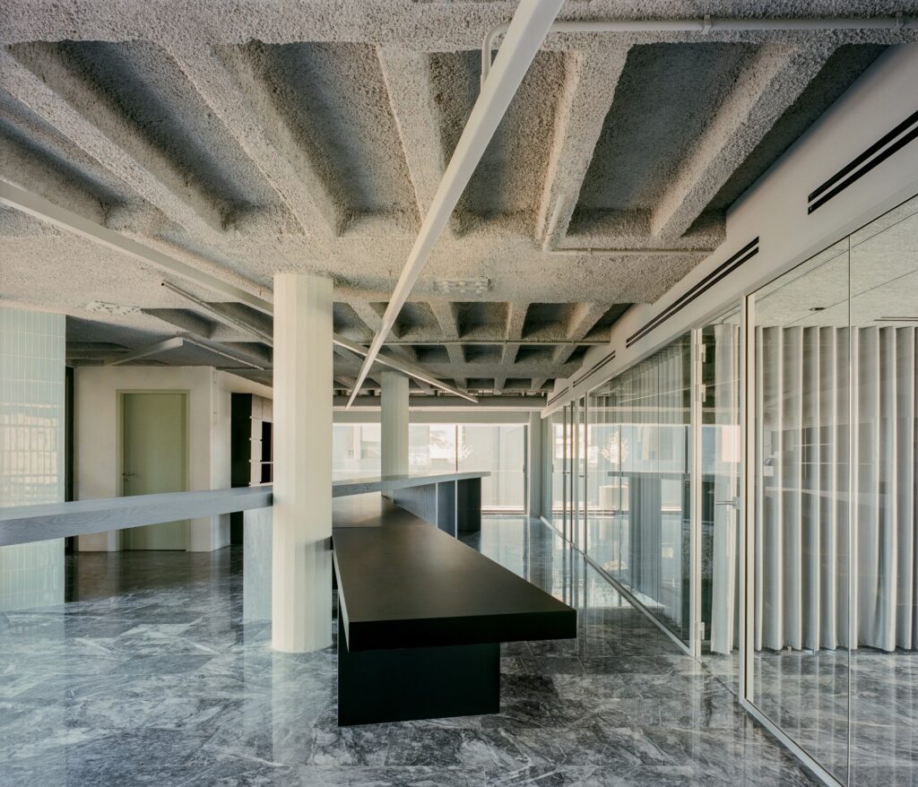 Modern office interior with an exposed concrete waffle slab ceiling, polished marble floors, and glass office partitions.